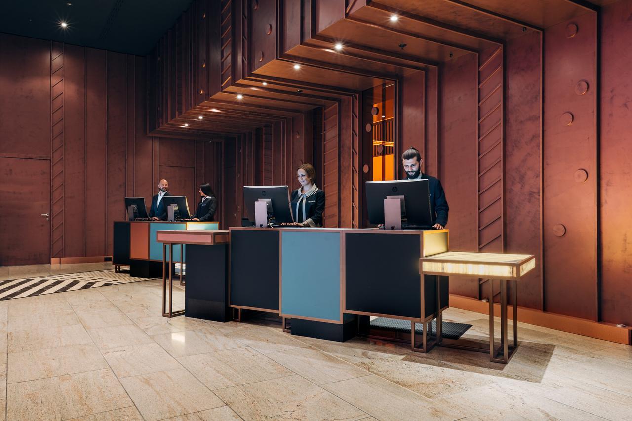 Hotel Pullman Berlin Schweizerhof Esterno foto The image shows a modern hotel lobby reception area. There are several reception desks made of dark materials with illuminated accents. Behind the desks, three people are standing, likely staff members, engaged with computer screens. The lobby featur